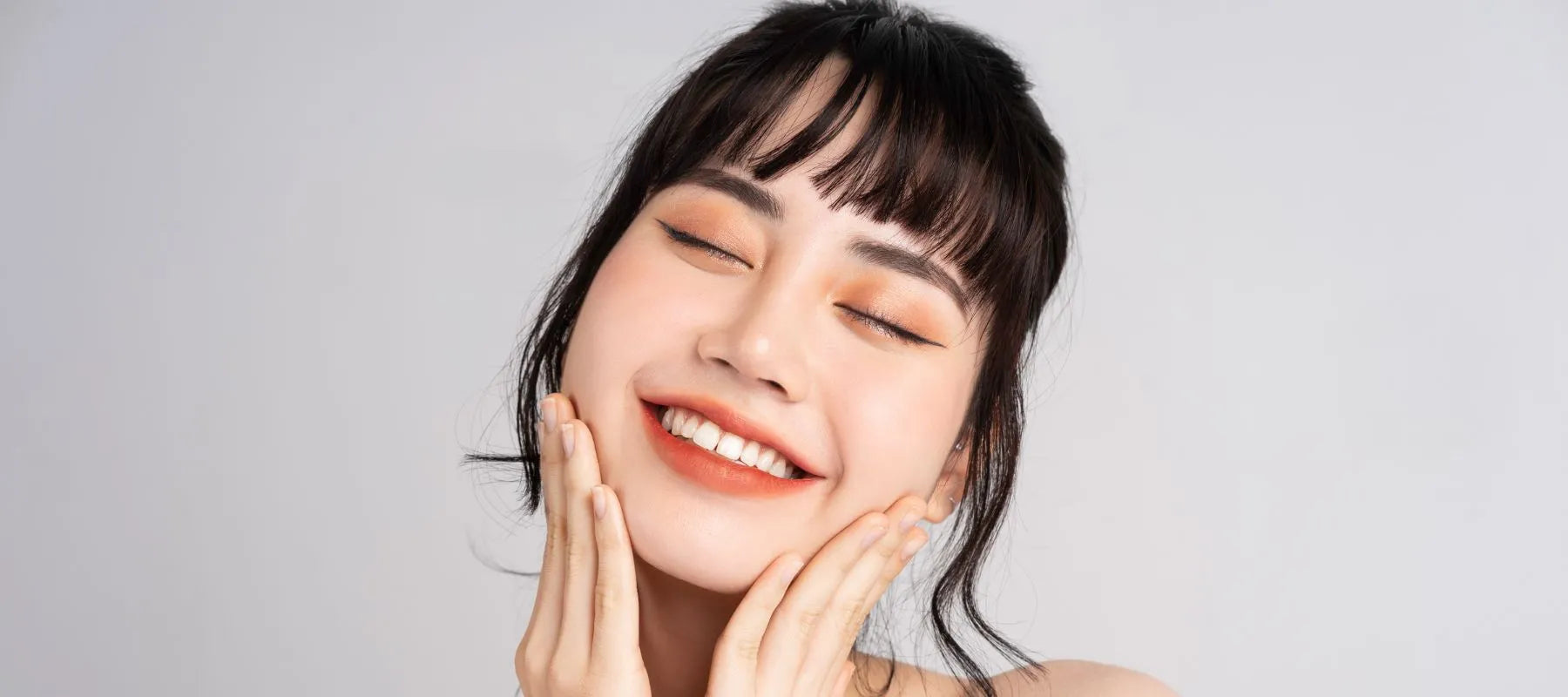 Close-up portrait of a joyful young Asian woman cradling her face, representing natural glowing skin and self-care